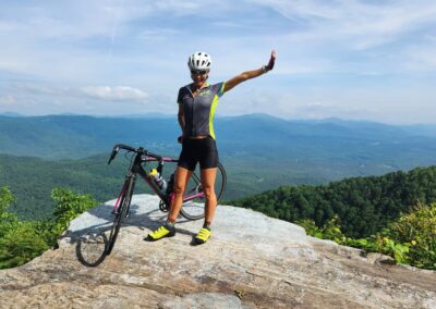 A Woman Standing On A Rock With A Bicycle And A Mountain View